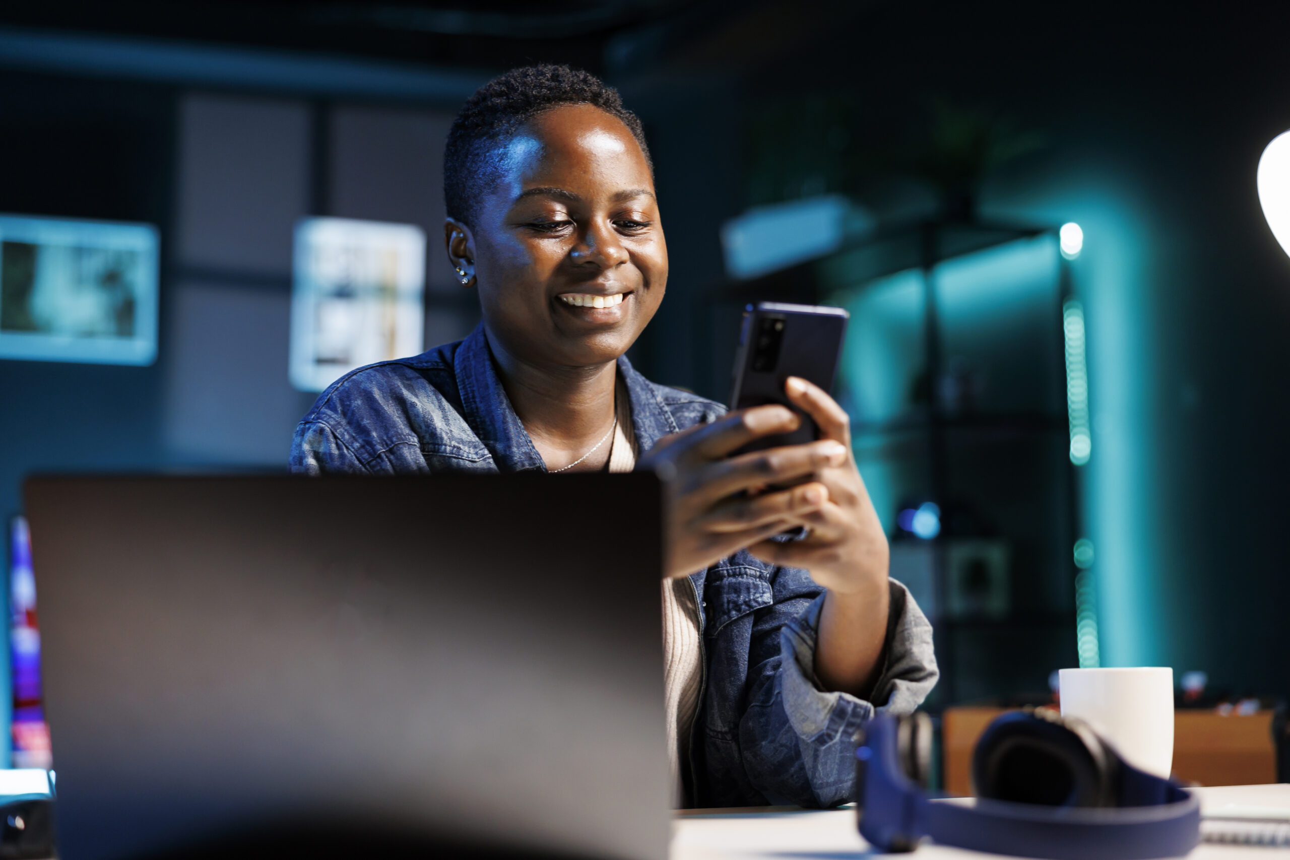 Cheerful female college student using mobile device to engage with friends on social media platforms. African american lady using cell phone to surf the web while sitting at desk in her home office.